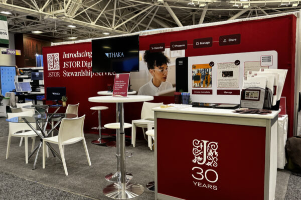A trade show booth for JSTOR’s Digital Stewardship Service, featuring red branding and a counter with a ‘30 Years’ logo. Behind the counter, a large backdrop displays visuals related to metadata creation and collection management. The booth includes a high table with stools, a glass table with chairs, two monitors showing the ITHAKA logo, and promotional materials neatly arranged on the counter.