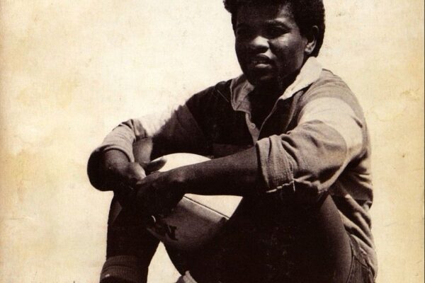 Cover of Common Lives/Lesbian Lives, a lesbian quarterly magazine. The title appears in red serif font at the top. Below the title, a Black woman in athletic clothing sits on grass, resting her arms on a rugby ball. She wears a striped rugby shirt, dark pants, high socks, and cleats. The photo is sepia-toned. At the bottom, the issue number is labeled “fifteen/sixteen” in red.