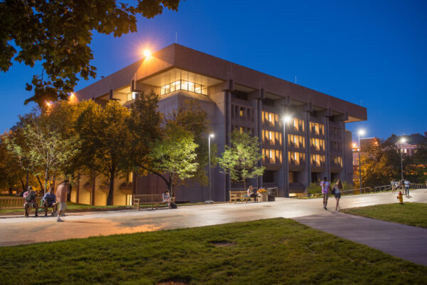 Bird Library at Syracuse University lit at night, with students walking and sitting on benches under glowing campus lights.