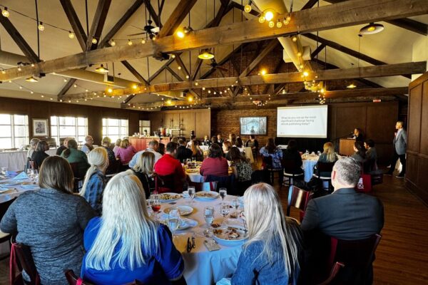 A group of attendees sits at round tables in a warmly lit event space with exposed wooden beams and string lights. A panel of four speakers is seated at the front behind a white table, with a presentation screen behind them. Many attendees have red tote bags hanging from their chairs with a white “J 30 Years” logo, celebrating JSTOR’s 30th anniversary.